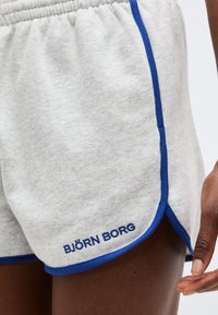 Lys grå atletiske shorts med blå kant og "BJÖRN BORG"-logo nær kanten, båret av en person med mørk hud, hånden synlig ved siden.