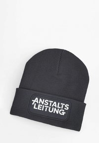 Black beanie hat with ribbed texture and a folded cuff featuring a rectangular patch with white text "ANSTALTSLEITUNG."