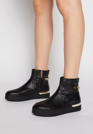Jambes portant des bottines noires avec motifs en relief, fermeture éclair dorée, détail chaîne et semelles épaisses noires sur un sol gris clair.