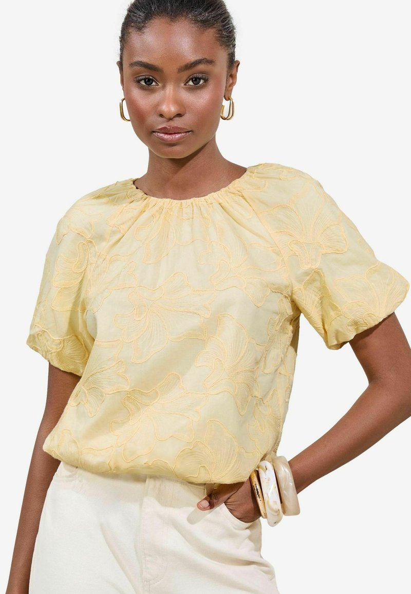 Geel geborduurde blouse met een geplooide halslijn en korte mouwen, met bloemenpatronen en lichte stof; gecombineerd met crèmekleurige broek.