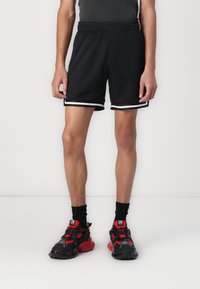Shorts de sport noirs avec une texture en maille, dotés de bandes latérales blanches. Associés à des baskets noires et rouges.