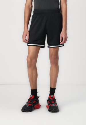 Shorts de sport noirs avec une texture en maille, dotés de bandes latérales blanches. Associés à des baskets noires et rouges.