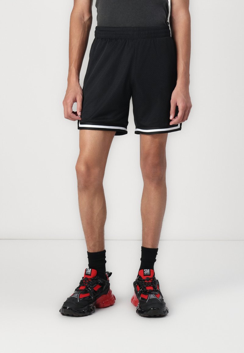 Shorts de sport noirs avec une texture en maille, dotés de bandes latérales blanches. Associés à des baskets noires et rouges.