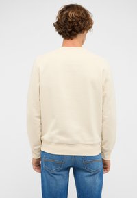 Beżowa bluza z długimi rękawami i ściągaczami przy mankietach. Gładka faktura, okrągły dekolt i czysty design, zestawiona z niebieskimi jeansami.