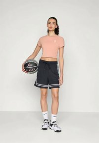 Ροζ κοντό τοπ, μαύρες μπασκετικές βερμούδες με λευκές ρίγες, λευκές κάλτσες και μαύρα αθλητικά παπούτσια. Κρατάει μια μπάλα μπάσκετ.