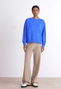 Blå sweatshirt med lange ermer, rund hals og ribbet kant, kombinert med beige vide bukser og hvite sko. Uformell antrekk.