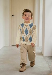 Crème trui met een blauw en bruin ruitpatroon, beige cargobroek en tan laarzen. Achtergrond bevat een lichtgekleurde deur en muren.