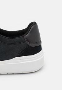 Svart sneaker med en texturerad mockaöverdel, slät läderhælaccent och en vit gummiyttersula. Sömnadsdetaljer synliga.
