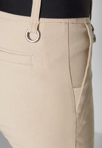 Beżowe spodnie wykonane z gładkiej tkaniny, z bocznymi kieszeniami, srebrnym metalowym pierścieniem i kontrastującym czarnym pasem w talii.
