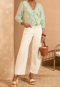 Kobieta w jasnozielonej bluzce z falbanami, białych, szerokich spodniach z wysokim stanem, na słomianych sandałach na koturnie, trzymająca małą brązową torebkę w kształcie wiadra.