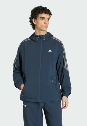 Junger Mann trägt eine marineblaue Adidas-Jacke mit Kapuze und drei grauen Streifen an den Ärmeln, steht mit teilweise in die Taschen gesteckten Händen.