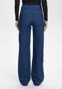 Tamsiai mėlyni plačiakojai džinsai, pasižymintys aukštu liemeniu, dviem užpakalinėmis kišenėmis ir švaria, lygiu tekstūra su subtiliais siuvinėjimo akcentais.