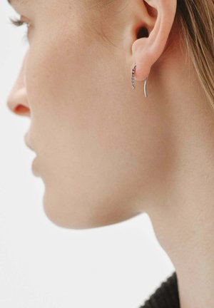 Pendientes de aro de plata con un diseño curvado y una fila de pequeñas piedras, mostrados en una oreja sobre un fondo neutro.