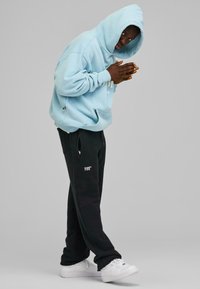Lys blå hettegenser med en stor frontlomme, kombinert med svarte joggebukser med sidelommer og en hvit logo på benet. Hvite sneakers.