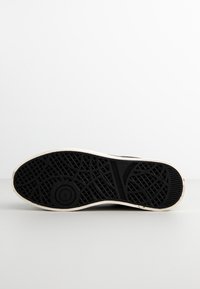 Semelle d'une chaussure en caoutchouc noir avec des motifs de bande de roulement géométriques et circulaires texturés, reposant sur une surface blanche avec un fond uni.