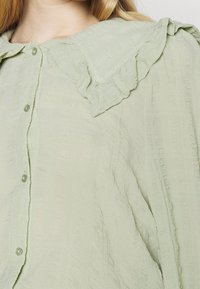 Light green blouse with a textured fabric, featuring a ruffled collar and button closure. Soft, flowing design with a relaxed fit.