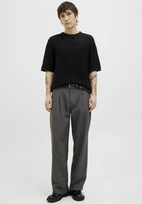 Black knitted short-sleeve sweater, gray pleated trousers, and black shoes. Tattoos visible on arms; textured fabric and a relaxed fit.