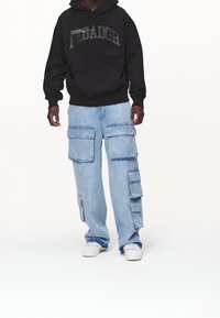 Felpa nera con cappuccio con la scritta "PECADOR", abbinata a pantaloni cargo in denim azzurro con numerose tasche grandi, indossata con sneakers bianche.