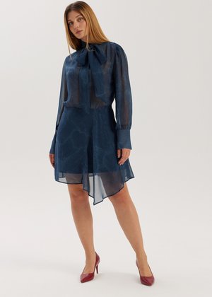 Przezroczysta granatowa bluzka z wiązaniem przy szyi i długimi rękawami, w połączeniu z pasującą spódnicą do kolan o asymetrycznym wykończeniu i miękkiej fakturze.