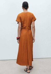 Rust-colored, ankle-length dress with short sleeves, a V-neck, and a back seam detail. Worn with a red and black bangle and sandals.