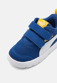 Zapatilla deportiva de malla azul con acentos blancos, que presenta dos correas de velcro y una etiqueta de logo amarilla en la lengüeta. Diseño de suela flexible.