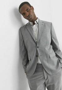 Šviesiai pilkas kostiumas su pritaikiu švarku ir derančiomis kelnėmis. Po juo stipriai juostuota marškiniai su mygtukais ir išryškintu apykakle. Lygi tekstūra.