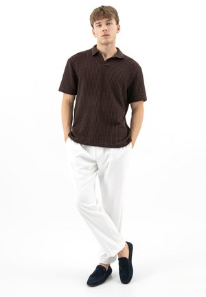 Young man standing with hands in pockets, wearing a dark brown textured polo, white pants, and black loafers on a white background.