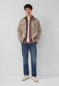 Beige Jacke mit Knopfleiste und zwei Fronttaschen, darüber ein rotes kariertes Hemd, weißes T-Shirt und blaue Jeans, kombiniert mit cremefarbenen Sneakern.