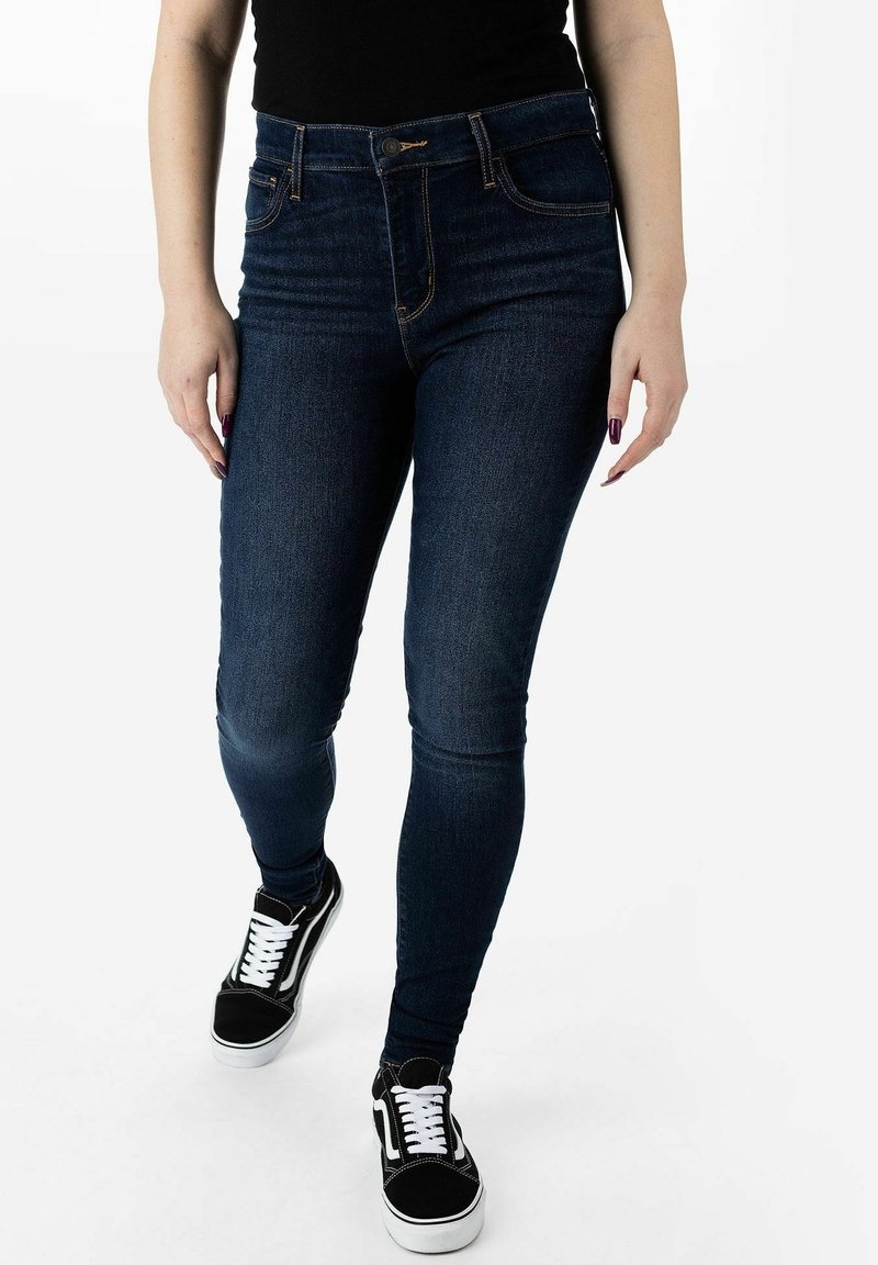 Jeans skinnies de tiro alto en azul oscuro con diseño clásico de cinco bolsillos, costuras en contraste y un aspecto desgastado; combinados con zapatillas negras.