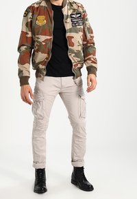 Maskádková bomber bunda s nášivkami, černé tričko, béžové cargo kalhoty a černé šněrovací boty. Bunda má elastické manžety a pas.