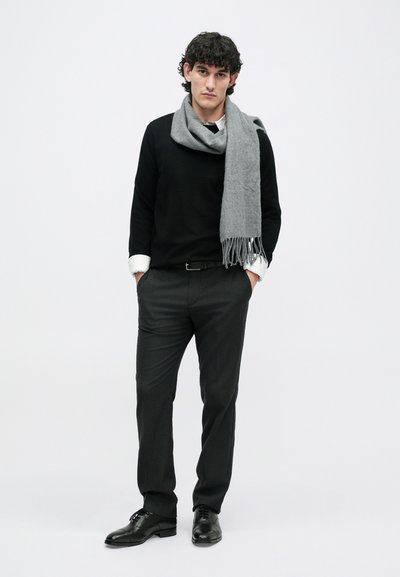 Pull noir, chemise col blanche, écharpe grise, pantalon sombre, chaussures noires ; modèle se tenant avec les mains dans les poches devant un fond uni.