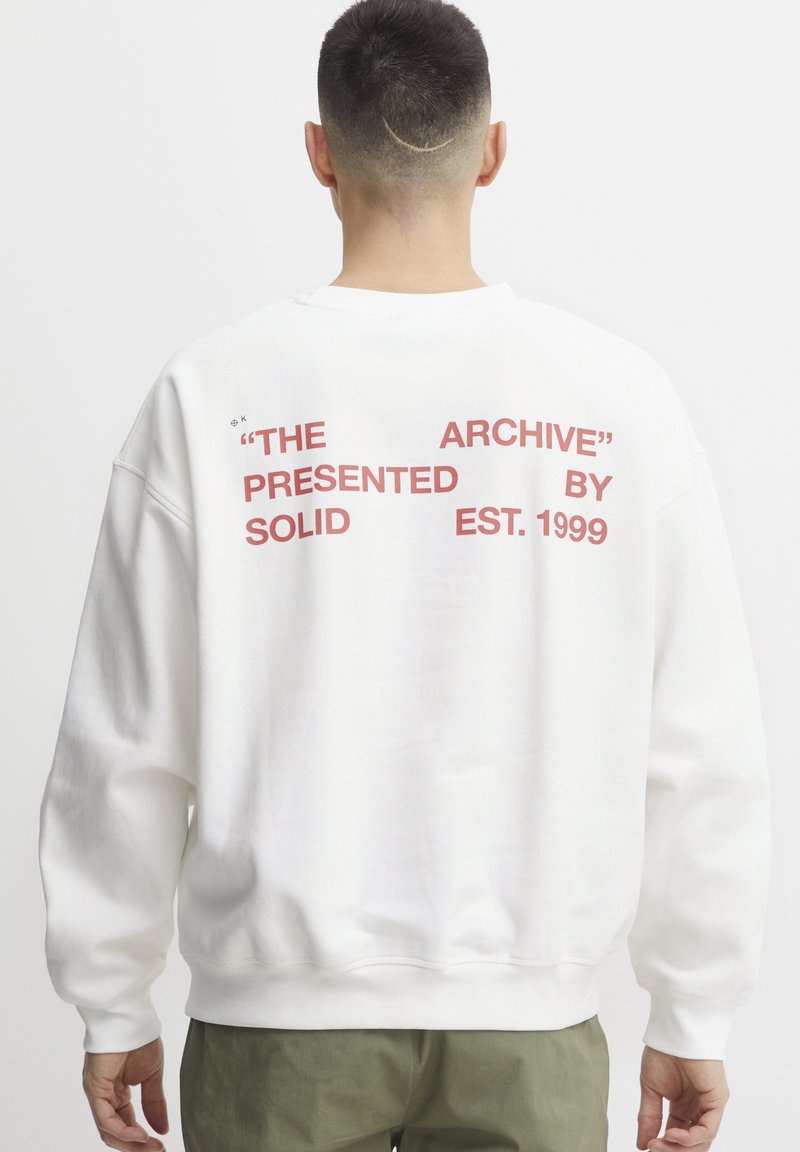 Sudadera blanca de corte oversize, con texto impreso en rojo en la parte trasera: "EL ARCHIVO" "PRESENTADO POR SOLID EST. 1999." Material de algodón suave.