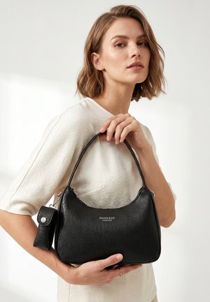 Woman in textured white top holding a black leather Diana & Co Firenze handbag with a small attached pouch against a plain background.