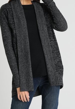Kvinde iført en mørkegrå cardigan med åbent front over en sort skjorte og blå jeans, med den ene hånd tæt på ansigtet og den anden ved siden af.