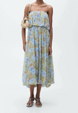 Vestido maxi floral en tonos verdes, azules y naranjas, con un cuerpo fruncido y tirantes ajustables. Combinado con un bolso crema.