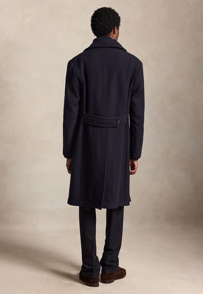 Man staat met de rug naar ons toe, draagt een lange donkerblauwe jas, donkere broek en bruine schoenen tegen een egale beige achtergrond.