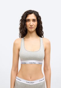 Γυναίκα με σγουρά σκούρα μαλλιά, φορώντας γκρι αθλητικό σουτιέν Calvin Klein και αντίστοιχο εσώρουχο, στέκεται μπροστά σε απλό λευκό φόντο.