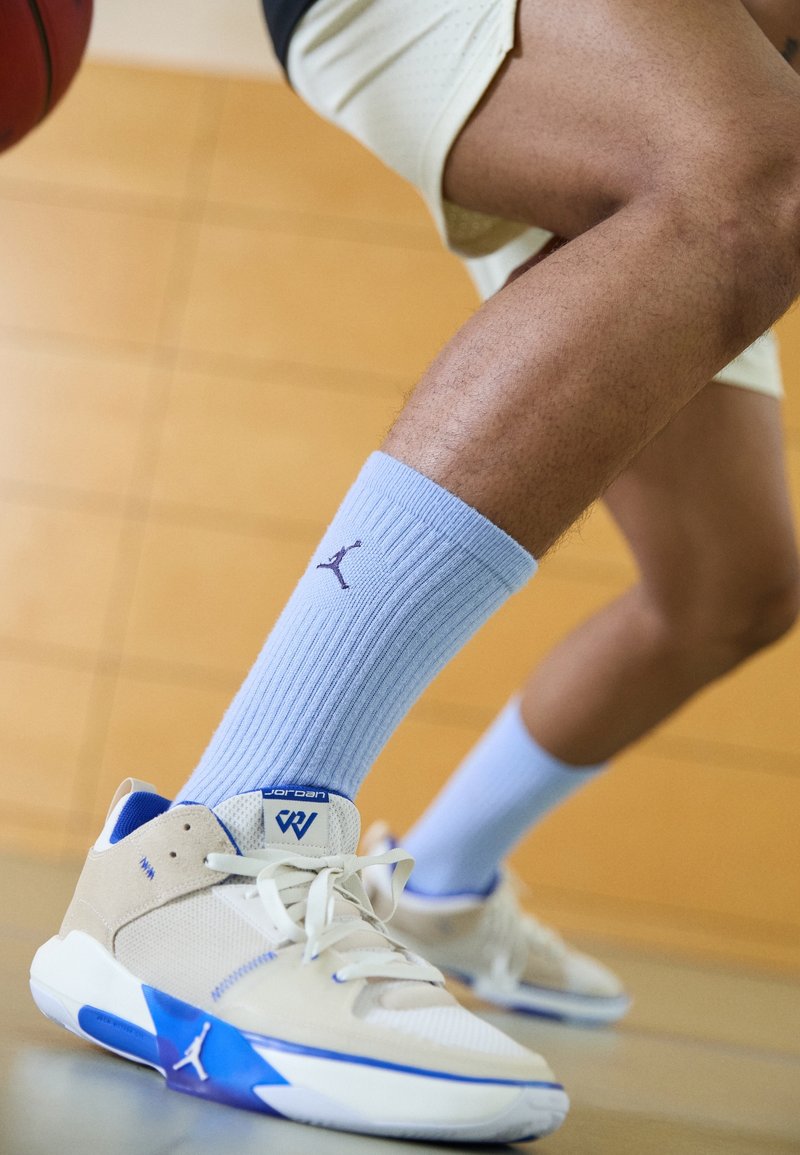 Chaussures de sport beiges avec des accents bleus, matériau texturé et semelle blanche. Chaussettes côtelées bleu clair avec un logo violet. Ballon de basket en arrière-plan.