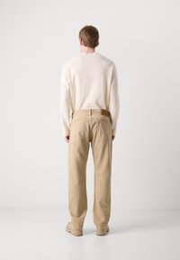 Pantaloni di cotone beige con gamba dritta e due tasche posteriori, abbinati a un maglione crema. Visti da dietro su uno sfondo semplice.