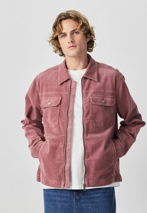 Junger Mann mit lockigem blondem Haar, trägt eine altrosafarbene Cordjacke über einem weißen Hemd und blauen Jeans, steht mit den Händen in den Taschen.