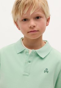 Polo shirt di un leggero verde menta con una finitura testurizzata, patta con tre bottoni e un piccolo logo di teschio e tibie sul lato sinistro del petto.