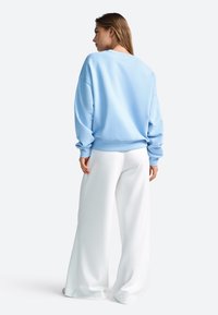 Gaiši zila džemperis ar vaļīgu piegriezumu, ribotām aprocēm un apakšiņu, kombinēts ar baltām platām biksēm, redzams no mugurpuses.