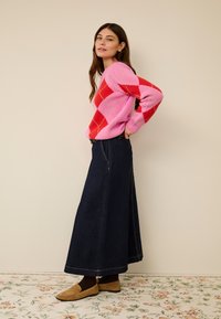 Różowo-czerwony sweter w szkocką kratę zestawiony z długą, ciemną spódnicą dżinsową. Strój ma luźny krój, subtelne przeszycia oraz buty w naturalnym kolorze.