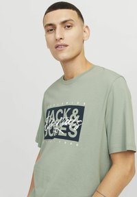 Zelené bavlněné tričko s černobílým grafickým potiskem s nápisem "JACK & JONES" umístěným uprostřed, kulatým výstřihem a krátkými rukávy.