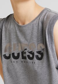 Camiseta sin mangas gris con el logotipo "GUESS" en texto negro y beige en capas. Hecha de una tela suave, con un cuello redondo y un corte casual.