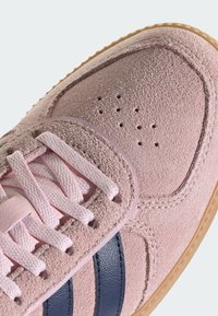 Chaussure de sport en suède rose avec semelle en caoutchouc crème, dotée de rayures bleu foncé, d'une tige texturée et de lacets rose clair.