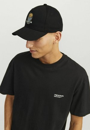 Gorra de béisbol negra con un gráfico bordado amarillo y gris, combinada con una camiseta de cuello redondo negra que presenta un pequeño logo blanco en el pecho.