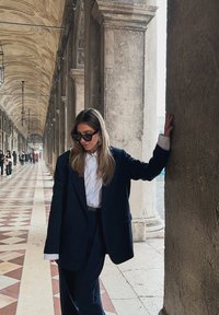 Juodas švarkas ir kelnės su balta marškinėliais. Blizgantys juodi saulės akiniai ir auksinės auskarai žiedai. Fone akmens kolonų ir raštuotas grindinys.