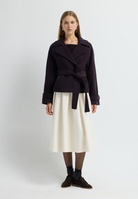 Lila omslagsoverall med breda slag och midjebälte, i kombination med en vit plisserad kjol. Bruna skor och texturerade strumpor fulländar looken.