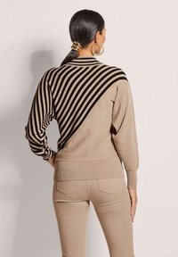 Strickpullover mit einem sandfarbenen Grundton und einem auffälligen schwarzen diagonalen Streifendesign. Gerippte Bündchen und Saum. Getragen mit beigen Hosen.
